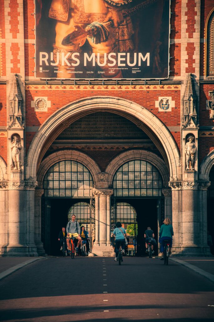 pexels-photo-12910498-12910498 Visitors cycle through the iconic arches of Amsterdam's Rijksmuseum, captured in a vertical frame.