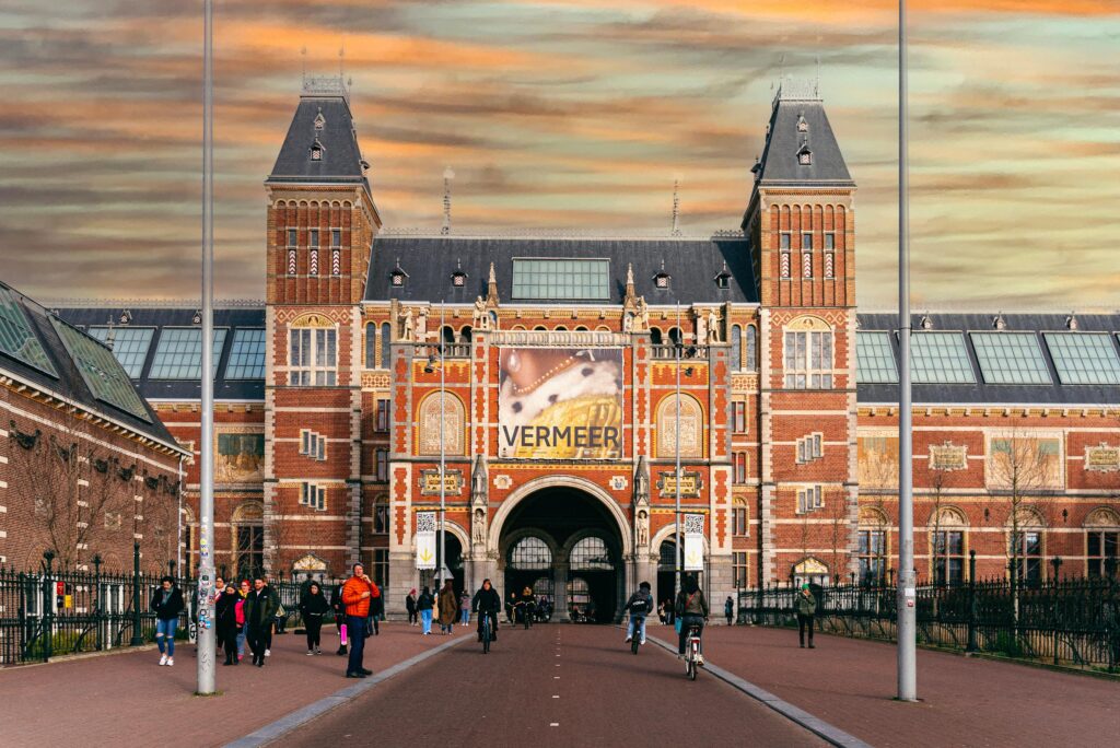 View of the iconic Rijksmuseum entrance in Amsterdam with people passing by during sunset.